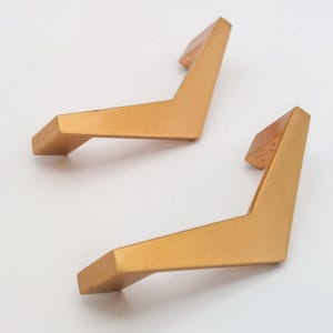 May include: Two gold-colored, V-shaped furniture handles. The handles have a rectangular base and a slightly angled, rectangular grip. The hardware is made of metal and has a matte finish. The handles are likely for a cabinet or drawer.
