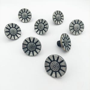 May include: A collection of eight round metal buttons featuring a sunburst pattern with a dark, recessed center and radiating lines. Each button has a brushed silver finish with black detailing in the grooves and a black shank on the back.