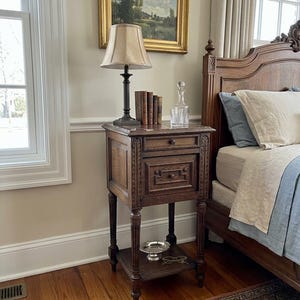 May include: A tall, dark wood nightstand featuring one small upper drawer and one larger lower drawer with decorative molding. The piece stands on four fluted legs and includes a lower shelf holding a small silver bowl and a pair of eyeglasses.