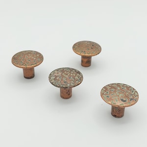 May include: Four copper-colored cabinet knobs with a textured, mottled surface. The knobs have a rounded, mushroom-like shape with a short, cylindrical stem. The surface has a mix of copper and green tones.