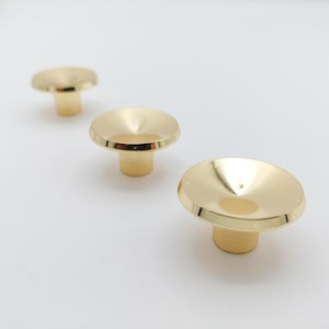 May include: Three gold-colored cabinet knobs with a rounded, disc-shaped top and a short, cylindrical base. The knobs are arranged in a diagonal line against a white background, showcasing their reflective surface and simple design.