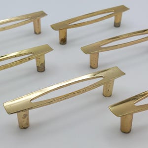 May include: A collection of six vintage brass cabinet pulls. Each pull features a rectangular handle with an oval cutout and two cylindrical posts. The brass has a warm, aged patina, suggesting the hardware's age and use. The pulls are arranged on a white surface.