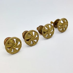 May include: Four antique brass cabinet knobs with a wheel-like design. The knobs have a warm, golden hue and are arranged on a white background. The decorative hardware features a detailed, openwork pattern, adding a vintage touch to furniture or cabinetry.