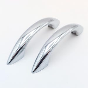 May include: Two shiny, chrome-finished door handles. The handles have a curved, ergonomic design, with a smooth, reflective surface. The handles are likely for a cabinet or drawer, and are set against a white background.