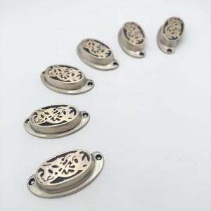 May include: A set of five oval-shaped metal drawer pulls with a brushed silver finish and a dark, intricate cutout pattern on the top surface. Each pull features two small screw holes on the side flanges for mounting to furniture or cabinets.