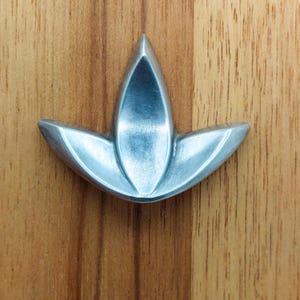 May include: A silver-toned metal cabinet knob featuring a three-petaled lotus or fleur-de-lis shape. The hardware has a smooth, brushed finish with curved, symmetrical edges that taper to a point at the top and sides, creating a clean look.