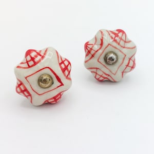 May include: Two decorative ceramic cabinet knobs. Each knob is white with a red geometric pattern and a central brass-colored screw. The knobs have a star-like shape with rounded edges, and are set against a white background.