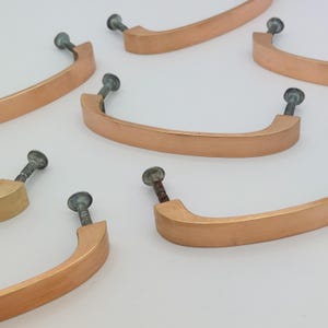 May include: Several copper-colored cabinet pulls with attached screws are arranged on a white surface. The pulls have a curved, rectangular design. The screws are silver and have a slightly aged appearance. The pulls are likely for furniture or cabinetry.