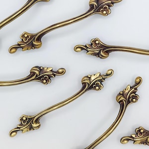 May include: Close-up of several antique brass drawer pulls. Each pull features an ornate floral design with curved handles and decorative ends. The pulls are arranged on a white background, showcasing their intricate details and warm, golden color.