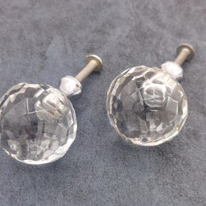 May include: Two clear, faceted crystal cabinet knobs with silver-colored metal hardware. The knobs are spherical and have a cut-glass appearance, reflecting light. They are set against a gray background.