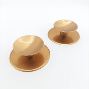 May include: Two gold-colored cabinet knobs with a circular base and a smaller, matching circular top. The knobs have a brushed metal finish and are set against a white background. These knobs are suitable for furniture.