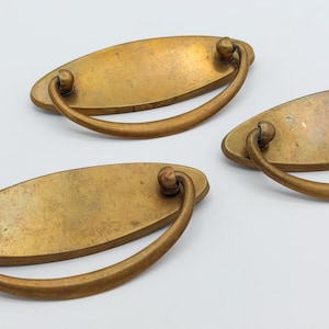 May include: Three antique brass drawer pulls with oval backplates and curved handles. The pulls have a warm, aged patina, suggesting vintage or reclaimed hardware. The handles are attached with small, round knobs.