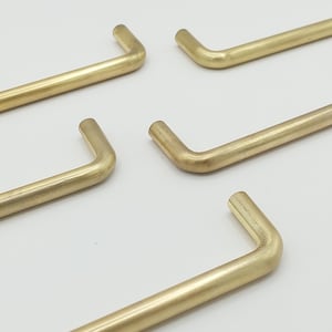 May include: Close-up of several brass-colored, L-shaped cabinet pulls. The pulls have a smooth, matte finish and are arranged against a white background. The design is simple and modern, suitable for various furniture styles.