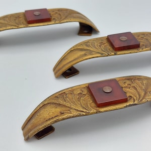 May include: Three antique-style cabinet pulls with a gold finish and ornate floral detailing. Each pull features a square, reddish-brown accent piece. The pulls are curved and have mounting hardware.