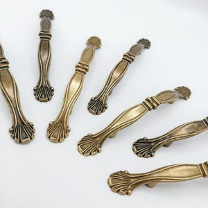 May include: A collection of six antique brass-toned metal drawer pulls. Each handle features a central elongated body with a smooth finish, flanked by decorative fan-shaped ends with vertical ridges. The hardware has a weathered, metallic patina.