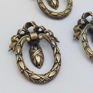 May include: Close-up of several antique brass drawer pulls. Each pull features a decorative oval ring with a leaf pattern, a bow at the top, and a central teardrop-shaped element. The pulls have a vintage, aged appearance.
