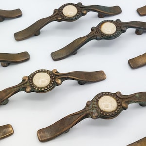 May include: Antique bronze cabinet pulls with a cream-colored center. The pulls have an ornate design with a curved handle and a decorative circular center. The hardware is likely for a cabinet or drawer.