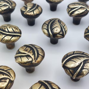 May include: A collection of antique-style cabinet knobs. Each knob features a detailed leaf design in a bronze finish. The knobs are round with a short stem, suitable for drawers or cabinet doors.