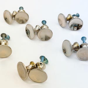May include: A collection of silver-toned cabinet knobs with a classic design. Each knob features a rounded, flat face and a decorative base, attached to a screw. The knobs are arranged on a white surface.