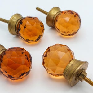 May include: Four amber-colored faceted glass cabinet knobs with brass hardware. The knobs have a geometric design and are mounted on threaded rods. The brass hardware includes a round base and a nut for securing the knob.
