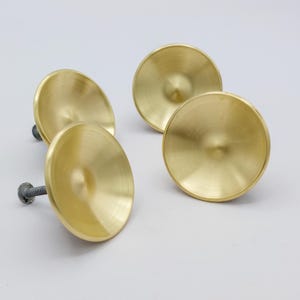 May include: Four brass cabinet knobs with a circular, disc-like design. Each knob has a central, conical protrusion and a threaded screw for mounting. The knobs have a polished, reflective surface and a warm, golden color.