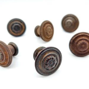 May include: A set of five circular cabinet knobs with a dark, weathered bronze finish. Each knob features a raised, concentric ring design with a central floral-like pattern and a beaded outer edge, showcasing a textured, aged metal appearance.