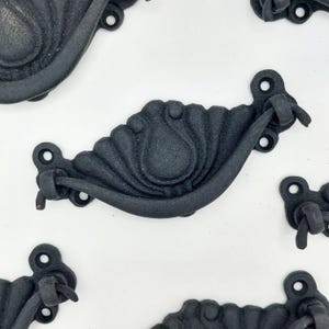 May include: Close-up of several black cast iron drawer pulls. Each pull has a decorative shell-like design and a curved handle. The pulls are arranged on a white surface, showcasing their intricate details and vintage style.