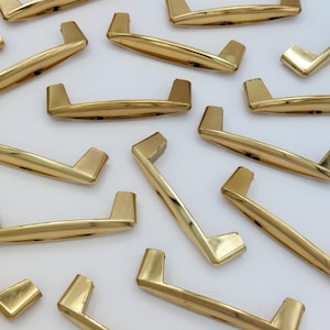 May include: A collection of gold-toned cabinet pulls with a sleek, modern design. The pulls have a rectangular bar with angled, upright posts on each end. The hardware is arranged on a white surface, showcasing their polished finish.