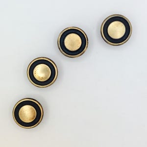 May include: Four circular knobs arranged in a diagonal pattern. Each knob features a central brushed brass-toned disc surrounded by a smooth, matte black circular ring, creating a distinct contrast between the metallic center and the dark border.
