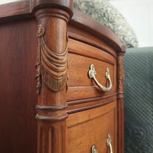 May include: Close-up of a wooden dresser with two drawers and ornate brass handles. The dresser has a dark brown finish with carved details on the corner post. The drawers have a curved front.