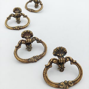 May include: Four brass-toned metal drawer pulls featuring a swinging oval handle attached to a decorative backplate with a shell-like motif at the top. The hardware displays a weathered, textured finish with intricate scrollwork details throughout.