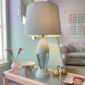 May include: A table lamp with a light blue ceramic base featuring a fluted design and a textured gray fabric drum shade. The lamp sits on a glass-topped table alongside a small pink vase, a patterned bowl, two brass swan figurines, and a magazine.