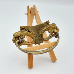 May include: Ornate antique brass drawer pull with a curved handle and decorative detailing. The pull is mounted on a small wooden easel against a white background. The handle is approximately 5 inches long.