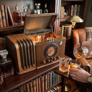 May include: A wooden liquor cabinet designed to resemble a vintage radio, featuring a hinged top lid that opens to reveal an illuminated interior storage area for bottles and glassware, with a circular speaker grille and control knobs on the front.