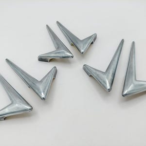 May include: A set of five silver-toned metal corner protectors, each featuring a sharp, V-shaped angular design with a smooth, reflective finish. These hardware pieces are arranged in a scattered pattern, showcasing their pointed ends and sturdy construction.