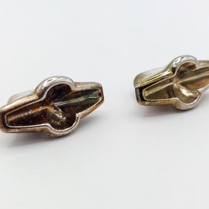 May include: A pair of silver-toned metal cufflinks featuring a stylized, elongated geometric shape with a central circular indentation and tapered ends. The metallic surface shows signs of wear and oxidation, with a polished finish on the edges.