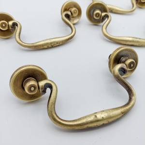 May include: Several antique brass drawer pulls with a curved handle design. Each pull has a circular backplate and a curved handle, all in a tarnished brass finish. The hardware is likely for furniture restoration or vintage decor.