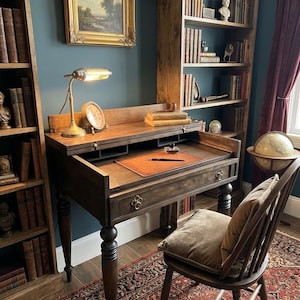 May include: A dark wood secretary desk with a hinged writing surface, a leather desk pad, an inkwell, and a fountain pen. The desk features turned legs and metal drawer pulls, accompanied by a wooden spindle-back chair with a velvet cushion.