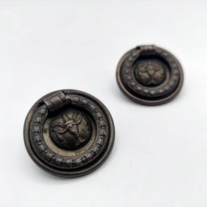May include: Two antique bronze drawer pulls with a circular design. Each pull features a central medallion with a classical figure, surrounded by a laurel wreath and a ring pull. The pulls are set against a white background.