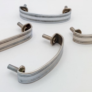 May include: Four vintage silver-toned cabinet handles. Each handle has a curved design with three horizontal lines and two mounting posts. The handles are arranged on a white surface, showcasing their design and potential for furniture restoration or DIY projects.