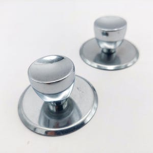 May include: Two shiny, chrome-colored cabinet knobs with a rounded top and a circular base. The knobs are set against a white background, reflecting light. The knobs are likely for drawers or cabinets.