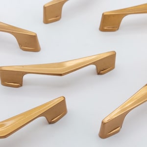May include: Several gold-colored cabinet handles are arranged on a white surface. The handles have a modern, angular design with a curved grip and rectangular mounting points. The finish appears to be matte.