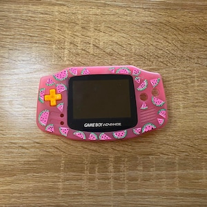 Puede incluir: Una consola de videojuegos portátil Game Boy Advance rosa con un diseño de sandía. La consola tiene una pantalla negra y un botón amarillo.