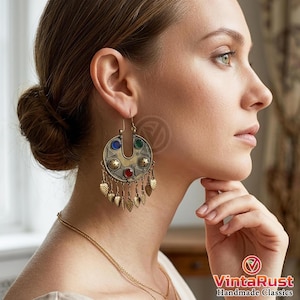 May include: A crescent-shaped brass earring featuring a central red stone, blue and green accents, and small circular metal details. The bottom edge is adorned with a fringe of dangling leaf-shaped charms that hang from the main frame.