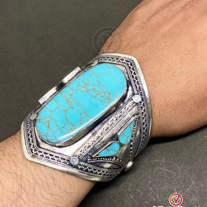 May include: A wide silver cuff bracelet featuring a large, oval turquoise stone. The bracelet has intricate metalwork with geometric patterns and smaller turquoise accents. The turquoise stone is a vibrant blue-green color.