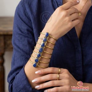 Könnte beinhalten: Ein goldfarbener Armreif mit sechs blauen Edelsteinen in einer vertikalen Reihe. Der Armreif hat mehrere horizontale Bänder und wird am Unterarm getragen. Goldringe und eine Halskette sind ebenfalls zu sehen. Der Hintergrund ist ein dunkelblaues Hemd.