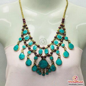 May include: A statement necklace featuring turquoise-colored stones and teardrop pendants. The necklace has a gold-tone chain and accents, with dark brown wooden beads. The necklace is displayed on a mannequin.