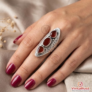 May include: A silver-colored ring with three oval, deep red gemstones. The ring has an elongated, ornate design with detailed patterns around the gemstones. The ring is on a hand with burgundy nail polish.