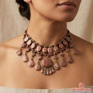 May include: A statement necklace featuring oval and teardrop-shaped pink gemstones with a mottled pattern. The necklace has a bronze-colored chain and accents, with a large central pendant and smaller dangles.