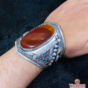 May include: A silver cuff bracelet with an oval, reddish-brown stone at its center. The bracelet features intricate silver detailing, including small turquoise and red accents. The design is ornate and the bracelet appears to be handmade.